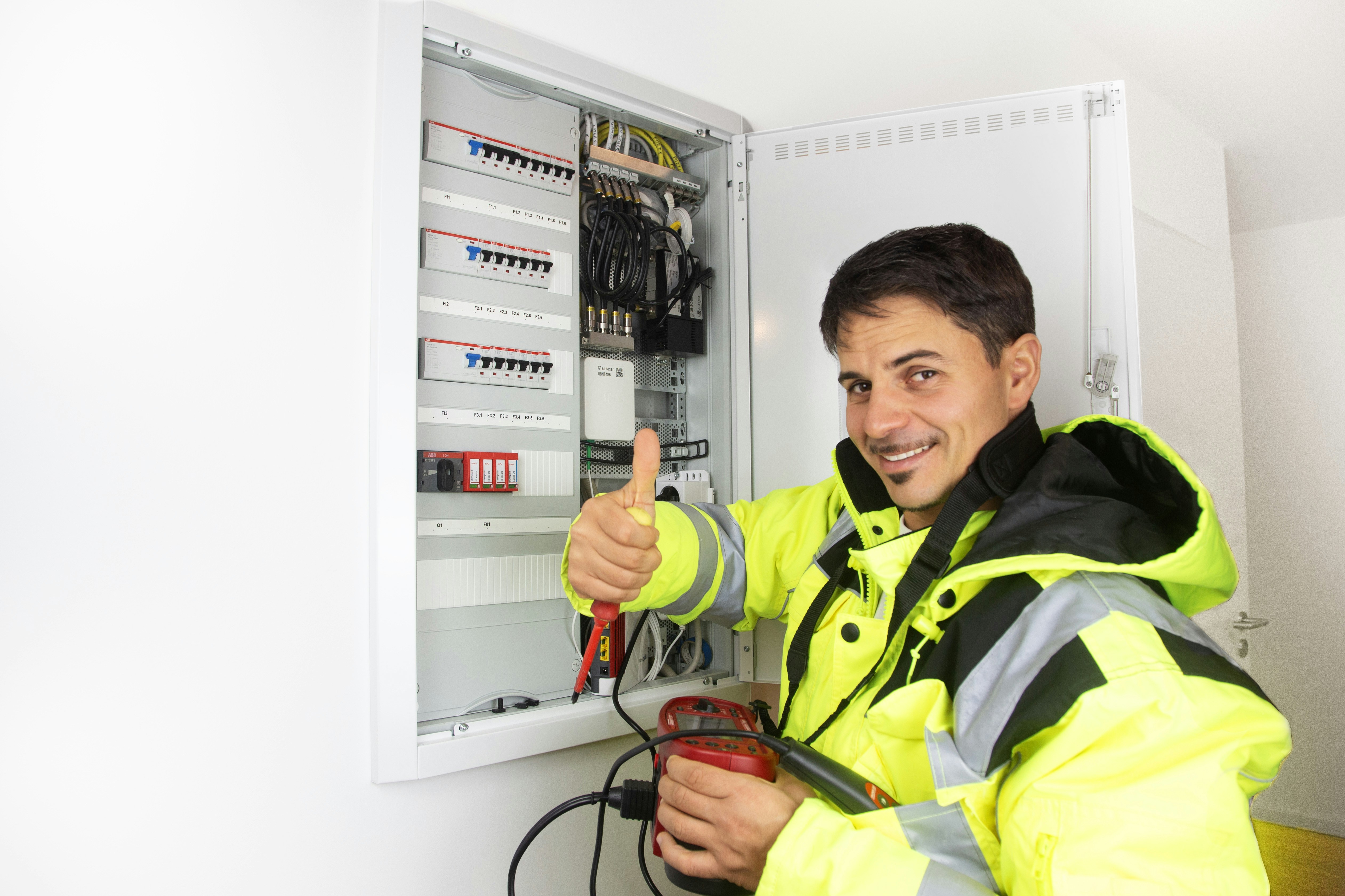 Ein männlicher Elektriker prüft den Zählerschrank, schaut in die Kamera und hält den Daumen nach oben.
